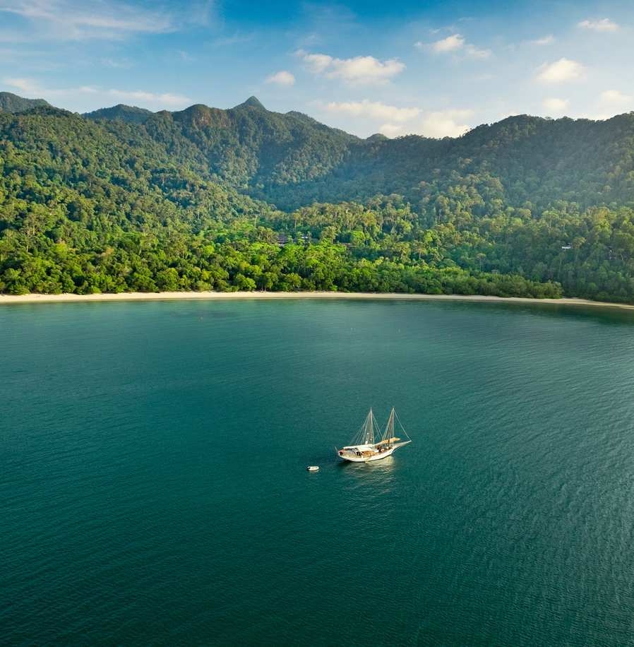 Aerial view of a sailboat anchored in a clear blue bay surrounded by high green mountains.