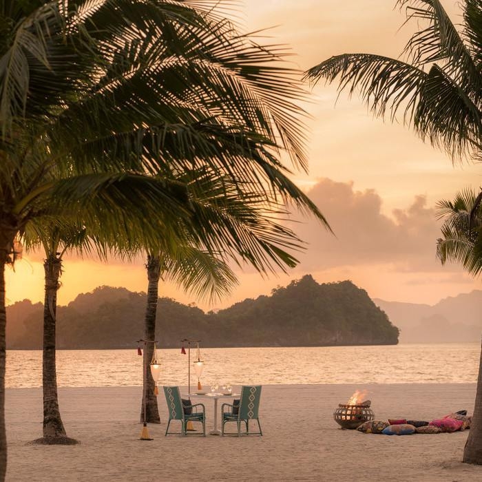 A romantic dinner table set for two on a sandy beach at sunset, flanked by palm trees and a small fire.