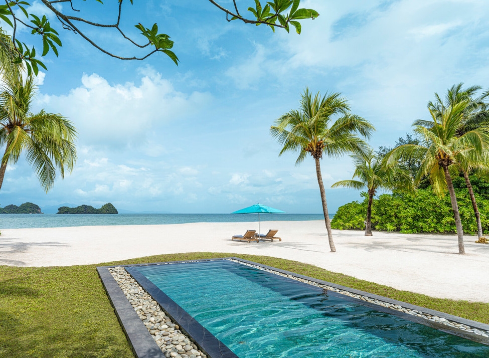 A private swimming pool leads to a white sand beach with turquoise water, palm trees, and a blue sun umbrella.