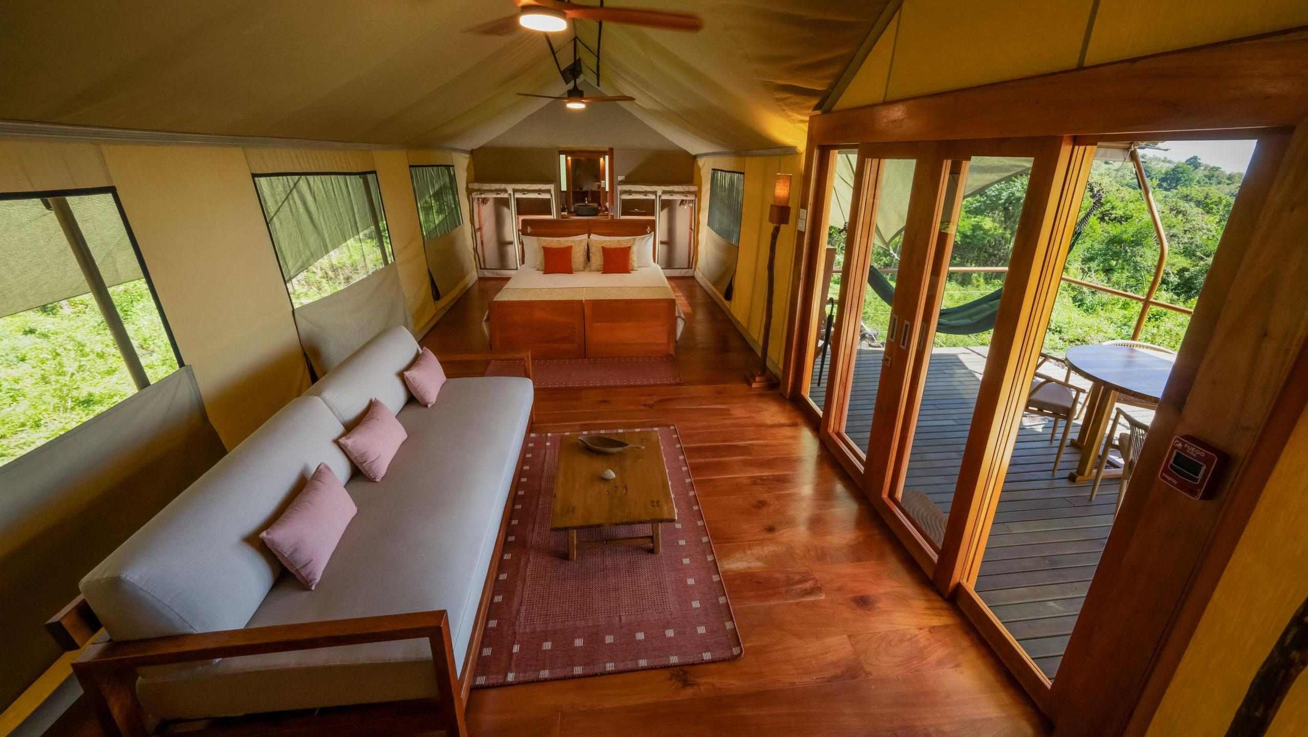 Interior of a canvas safari tent with a sofa, wooden floor, double bed in the background, and open deck glass doors.