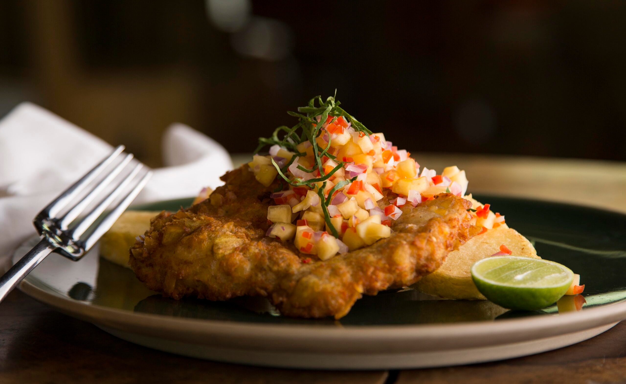 A breaded chicken cutlet topped with diced fruit salsa, served with lime and garnish.
