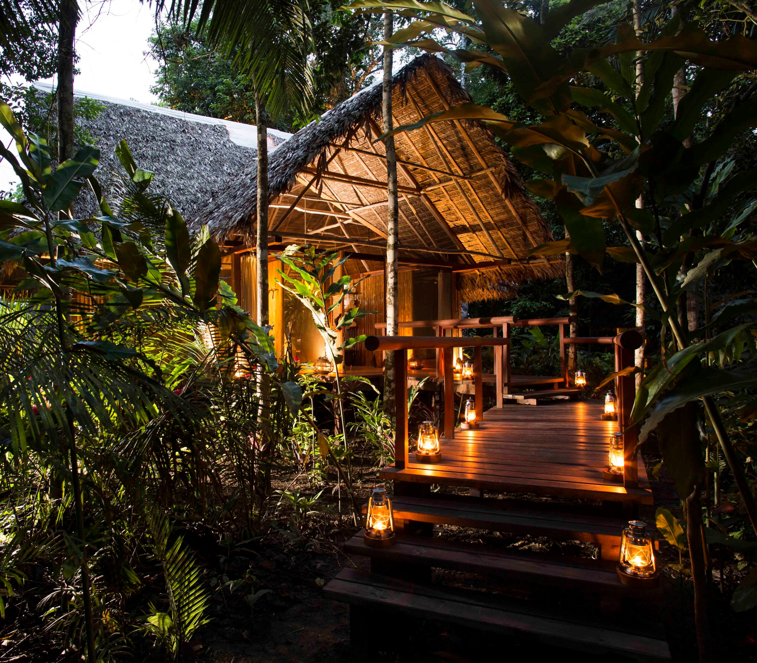 Thatched-roof cabin with lit lanterns on a wooden deck in a tropical forest at dusk.