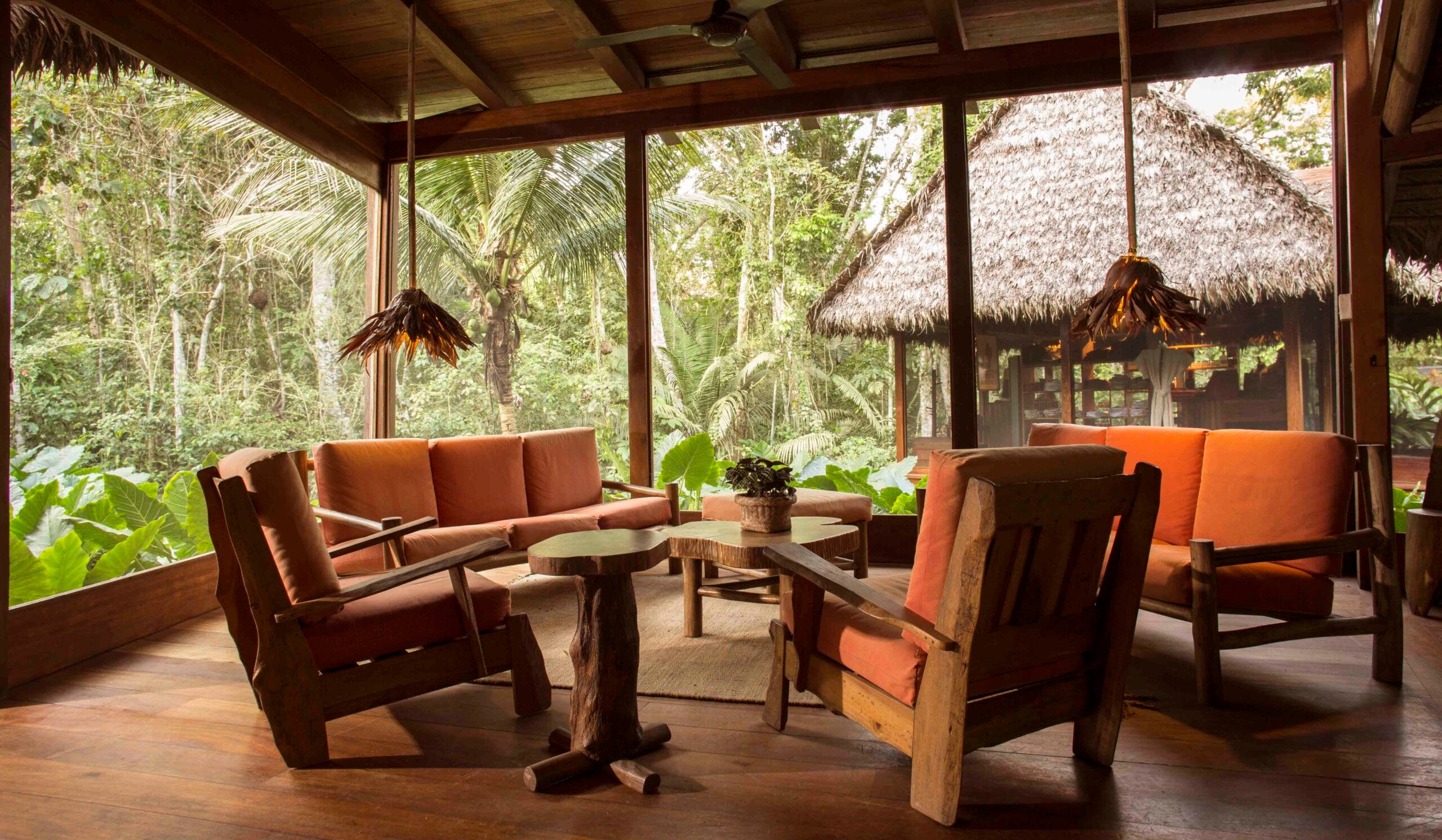Rustic lounge with wooden furniture and tropical forest view outside large windows.