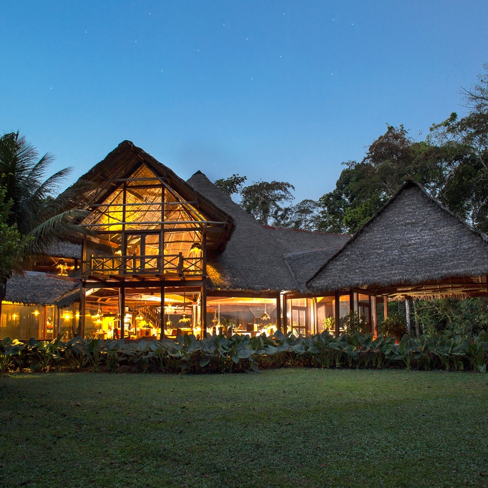 Illuminated lodge with thatched roofs at twilight surrounded by tropical vegetation.