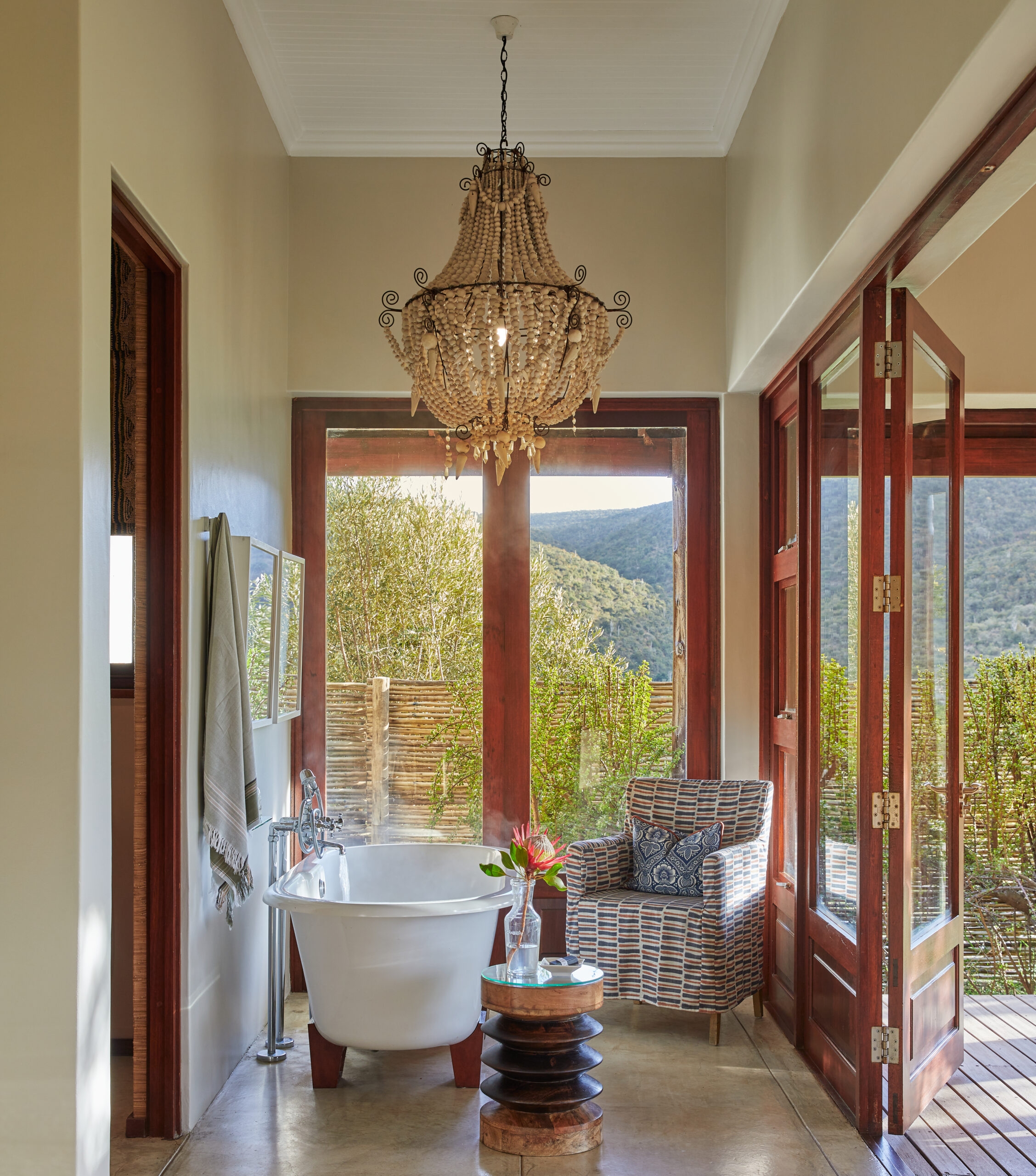 Luxurious bathroom with a freestanding tub and chandelier, opening to a scenic view of nature.