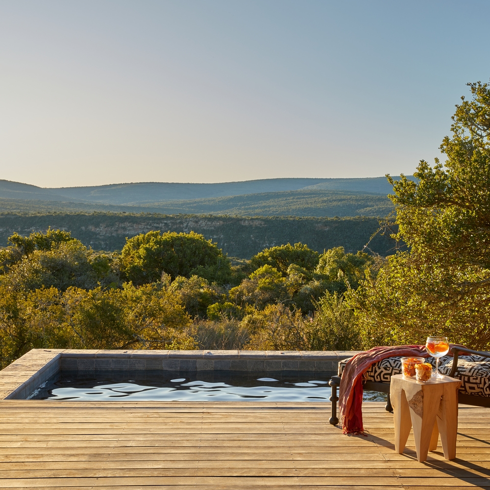 Wooden deck with a pool overlooking a forested valley at sunset, with drinks and snacks on a side table.