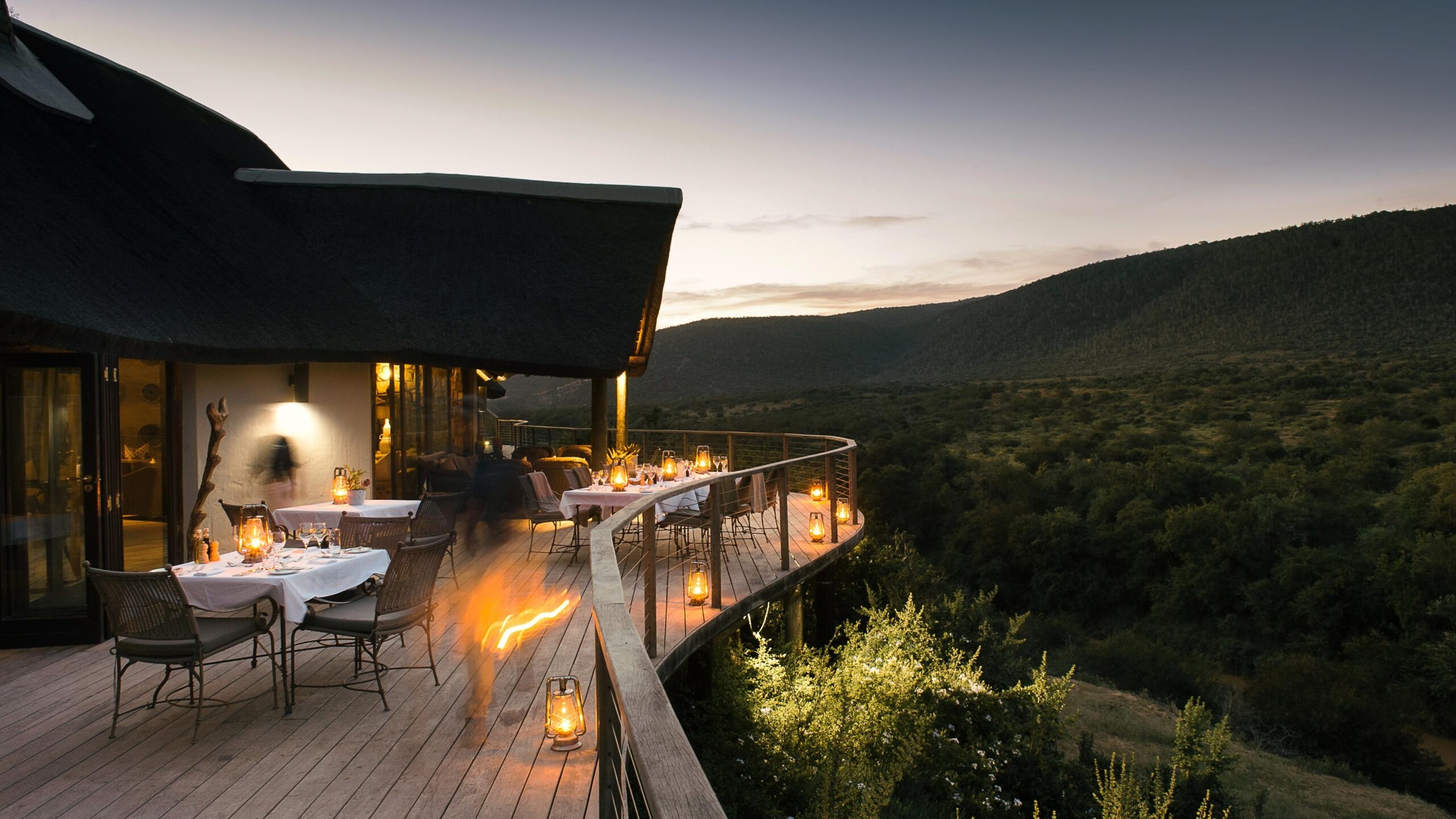 Terrace of a luxury lodge with dining setup overlooking a lush valley at dusk.