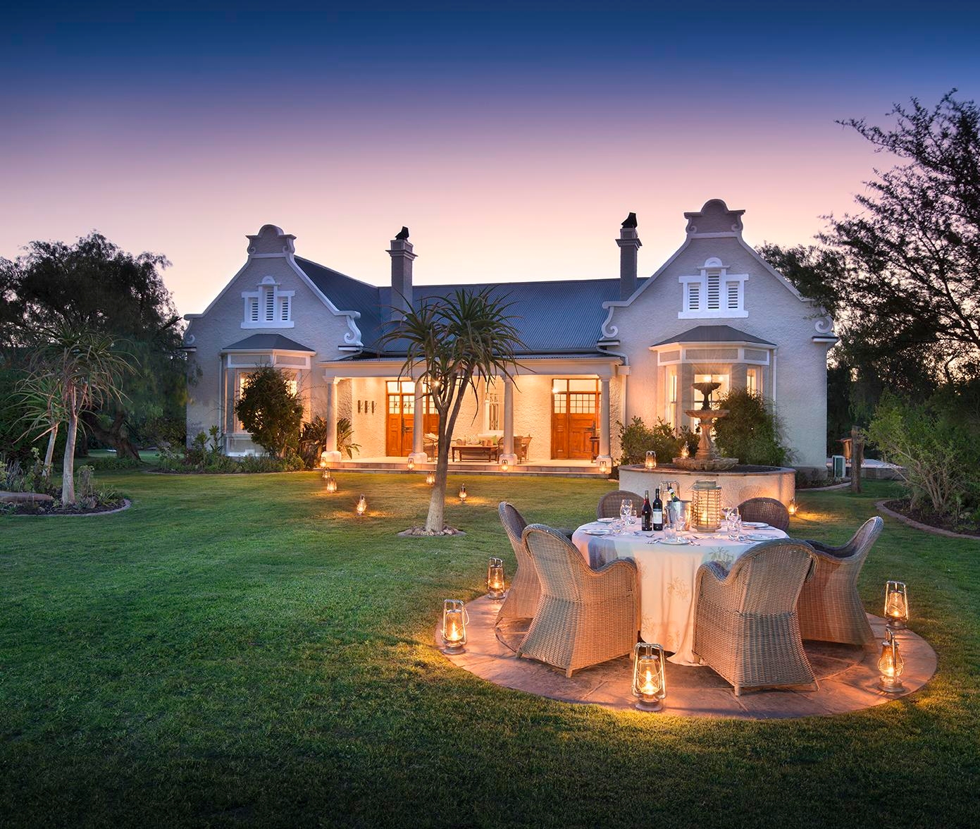 Elegant house with outdoor dining setup at dusk, surrounded by lit lanterns.