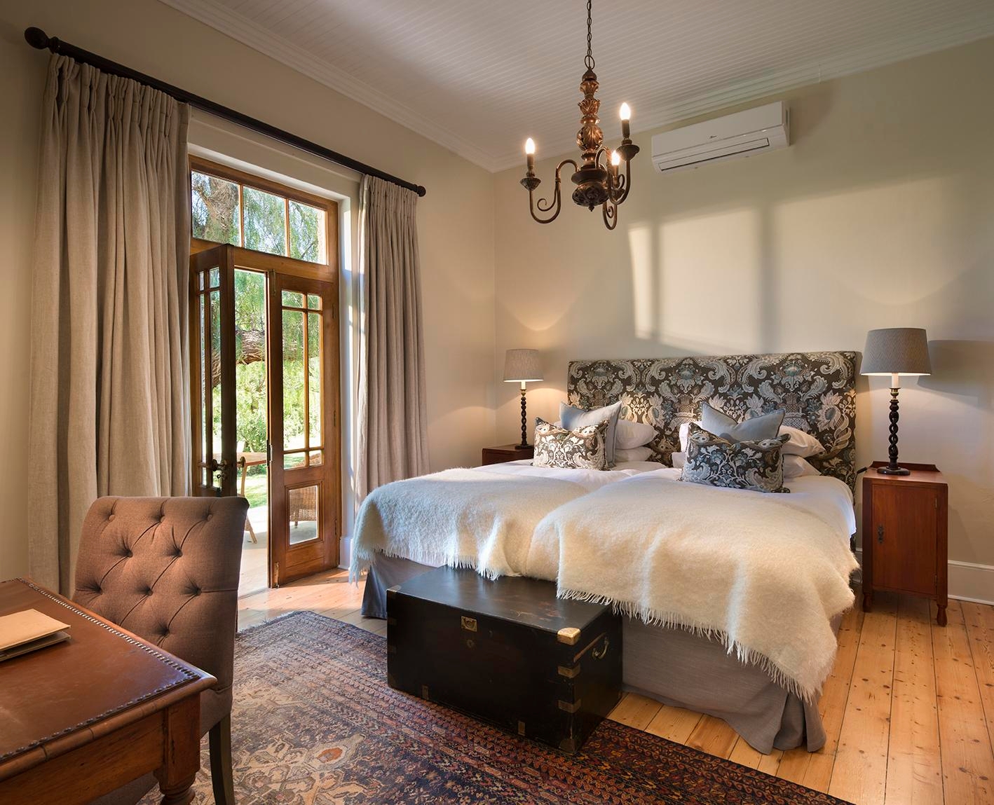 Elegant bedroom with twin beds, patterned headboard, chandelier, and French doors leading outside.