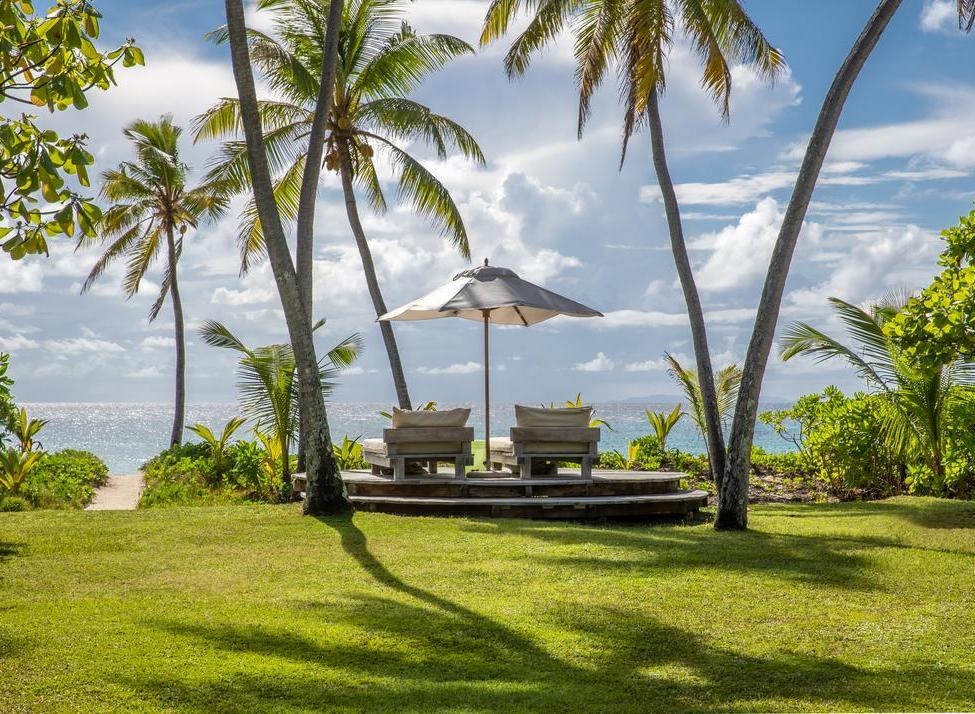 Beachfront lounge chairs under an umbrella on a wooden deck surrounded by palm trees and green grass.