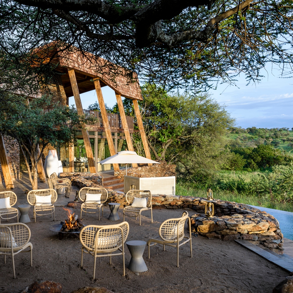 Modern seating around a fire pit near a stonewall house, surrounded by greenery and trees, creating a serene outdoor gathering space.