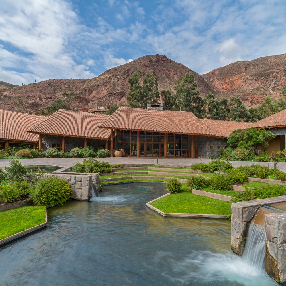 Rustic building with a tiled roof, manicured garden, and flowing water feature against a mountain backdrop.