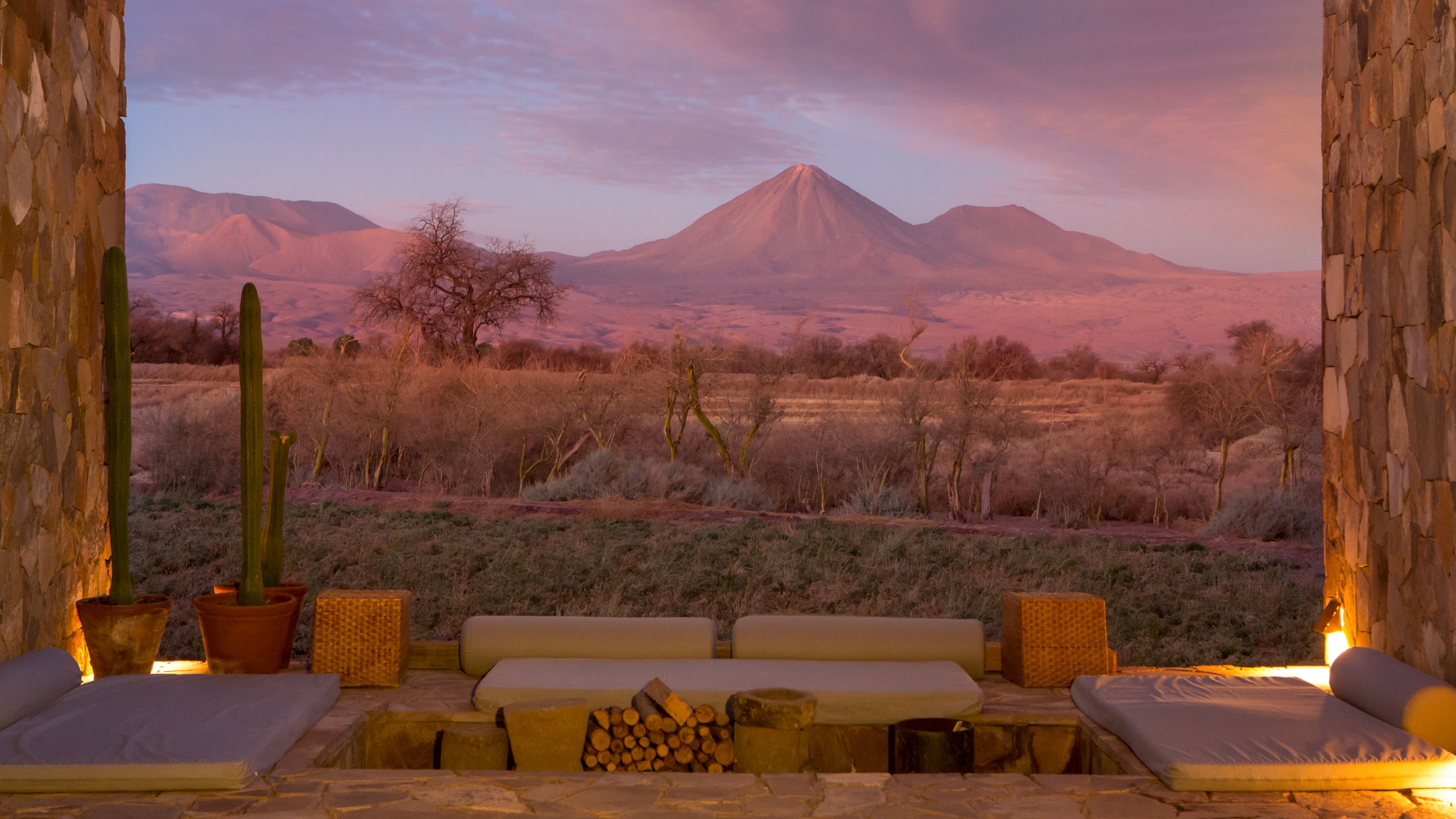 Sunken lounge with cushions and a fire pit framing a view of a large volcano and desert landscape at sunset.