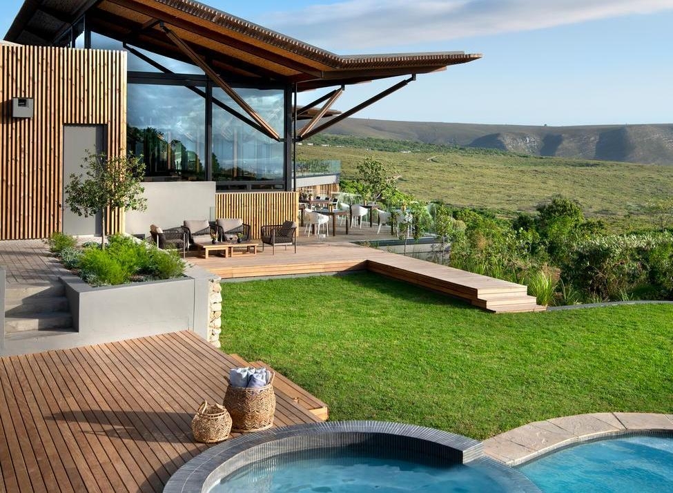 A modern glass-walled lodge with a wooden deck and outdoor seating overlooking a vast green valley under a blue sky.