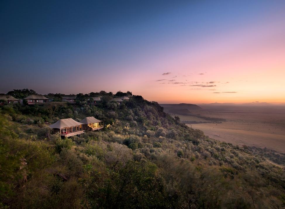 Luxury safari tents on a green hillside at sunset overlooking a wide African savannah landscape.