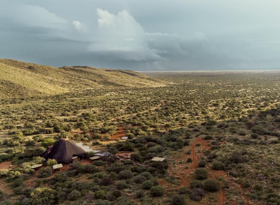 An aerial view of a thatched-roof lodge situated in a vast savanna landscape beneath cloudy skies.