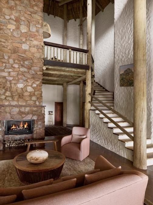A living room with a stone fireplace, plush armchairs, wooden columns, and a staircase leading upstairs.