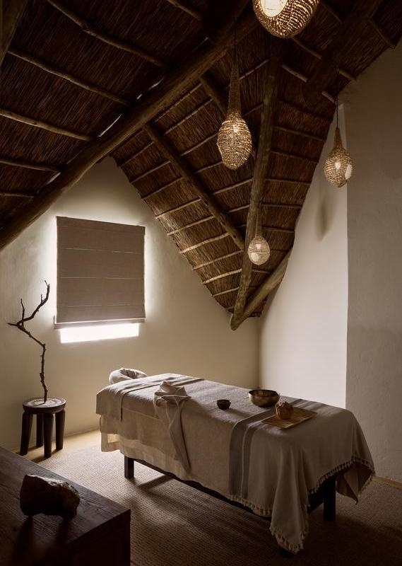 A serene spa treatment room with a covered massage bed under a thatched roof with woven pendant lamps.