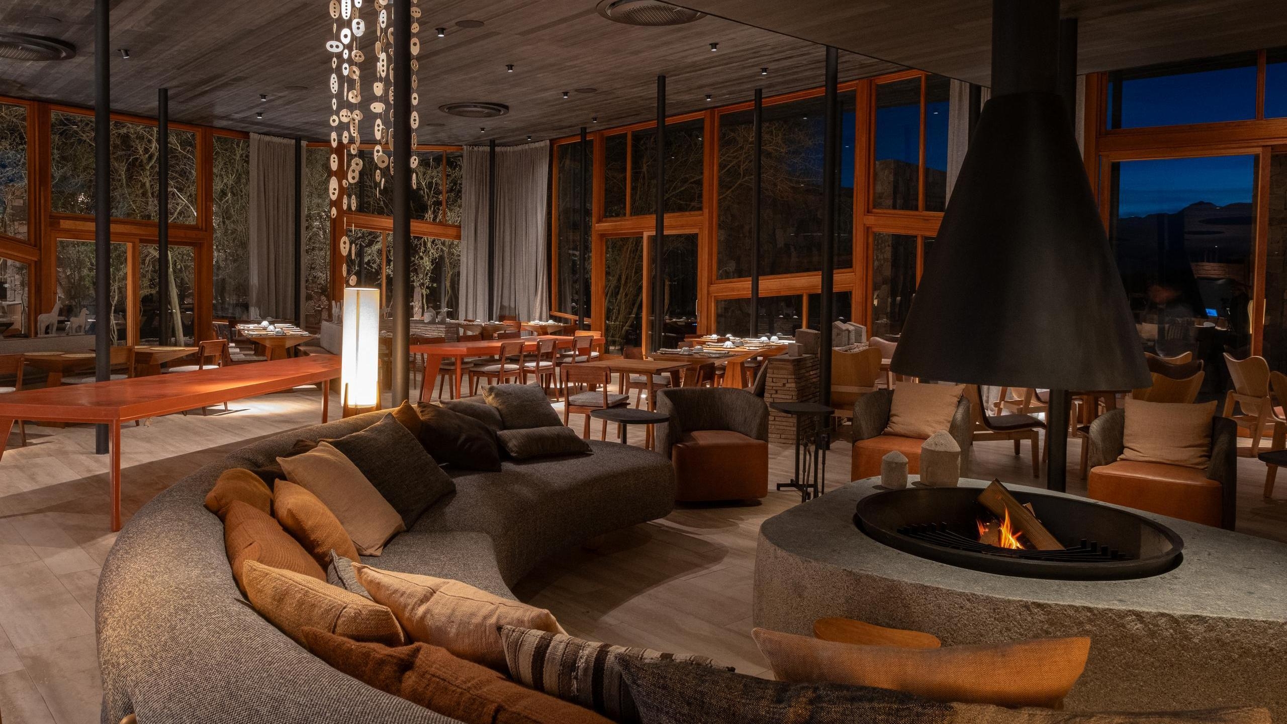 Cozy hotel lounge interior showing a curved gray couch, a large stone fireplace with burning wood, and dining tables.