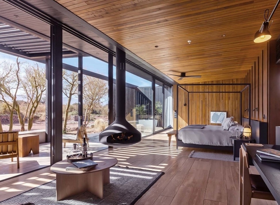A modern luxury hotel room with wood walls, a four-poster bed, and a suspended black fireplace by large glass doors.