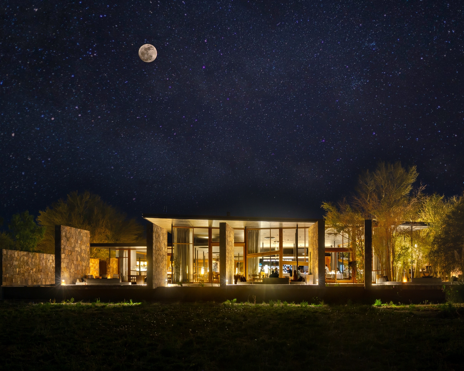 A modern stone and glass building illuminated at night under a clear starry sky featuring a bright full moon.