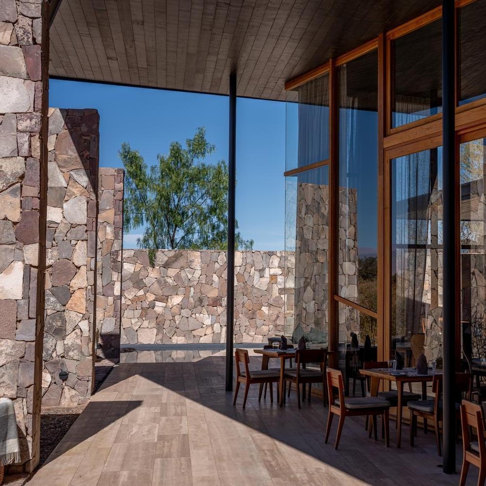 A sunny outdoor hotel terrace with wooden dining tables and chairs set against large, rustic stone accent walls.