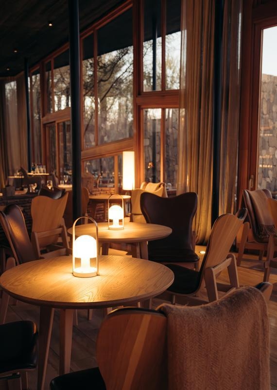Interior view of a hotel lounge featuring round wooden tables illuminated by glowing dome lamps and cozy chairs.