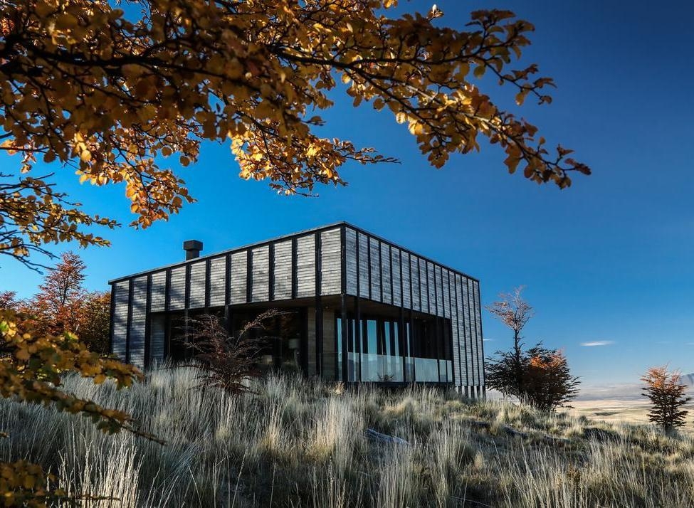 A small wooden building in a vast field with towering, snow-covered jagged mountains in the background.