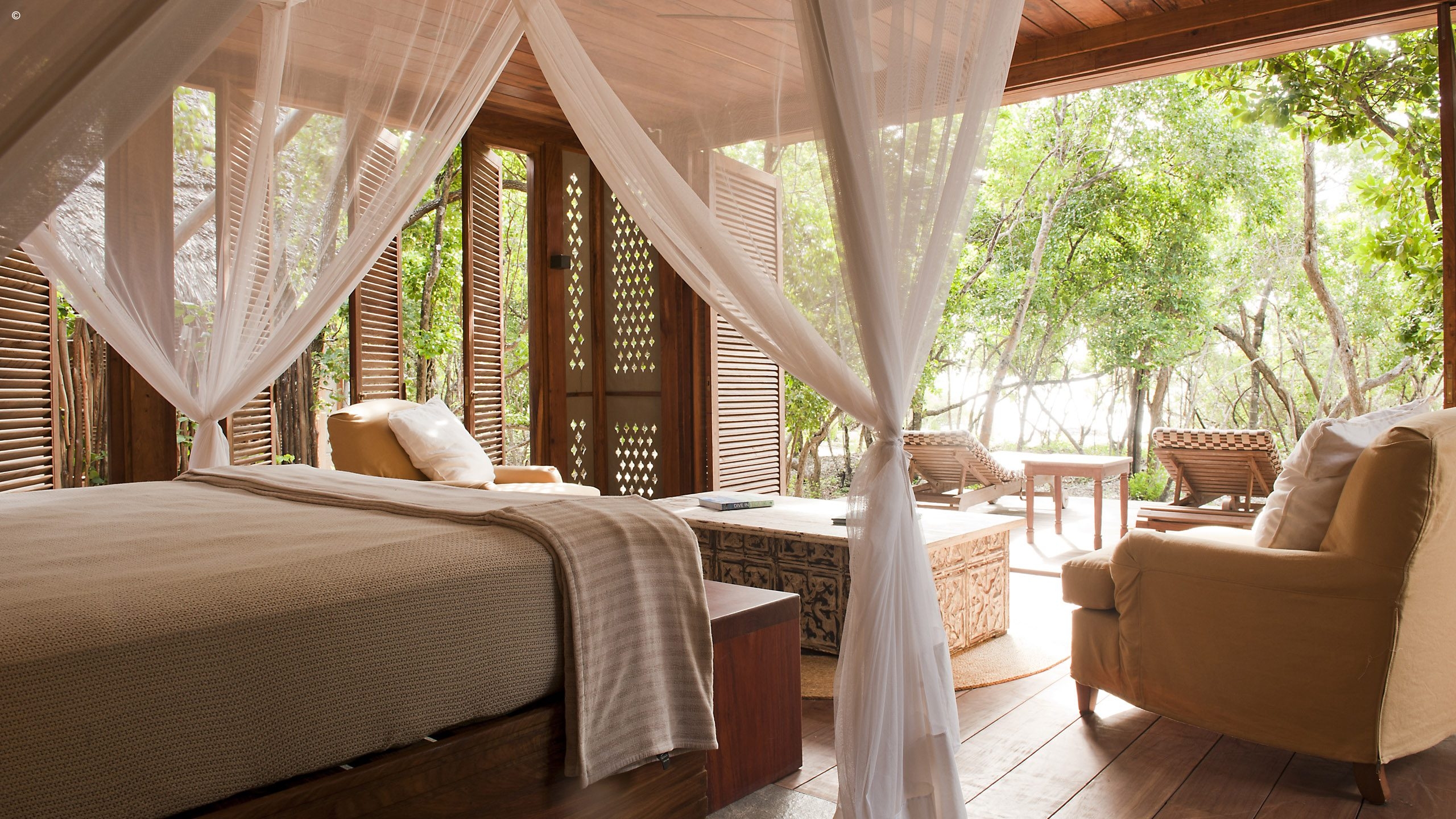 A serene, open-air bedroom with a canopy bed, wooden furnishings, and views of lush greenery outside. Natural light floods the space.