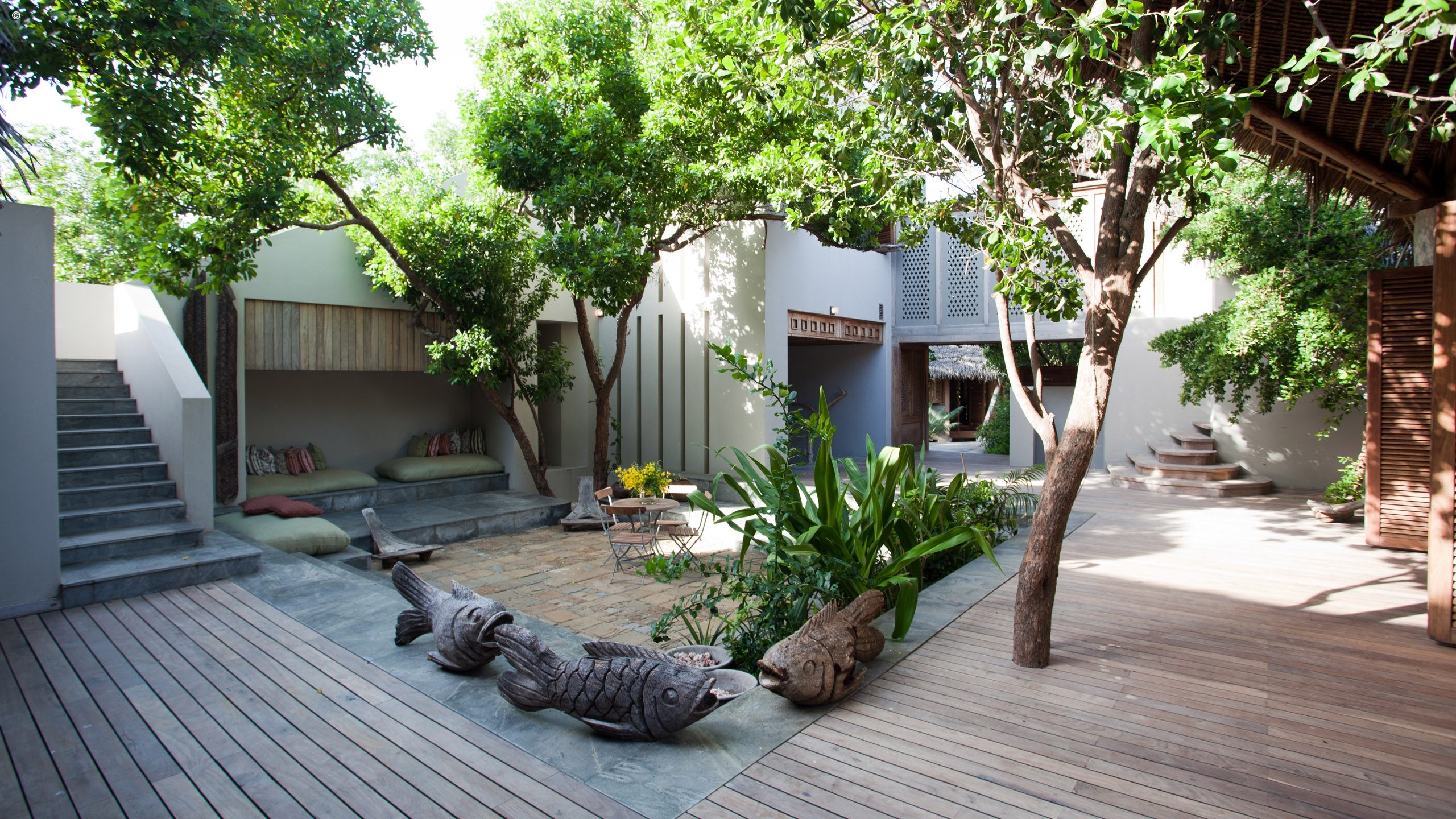 A serene courtyard features lush greenery, wooden steps, a stone pathway, and carved fish sculptures, creating a tranquil outdoor space.