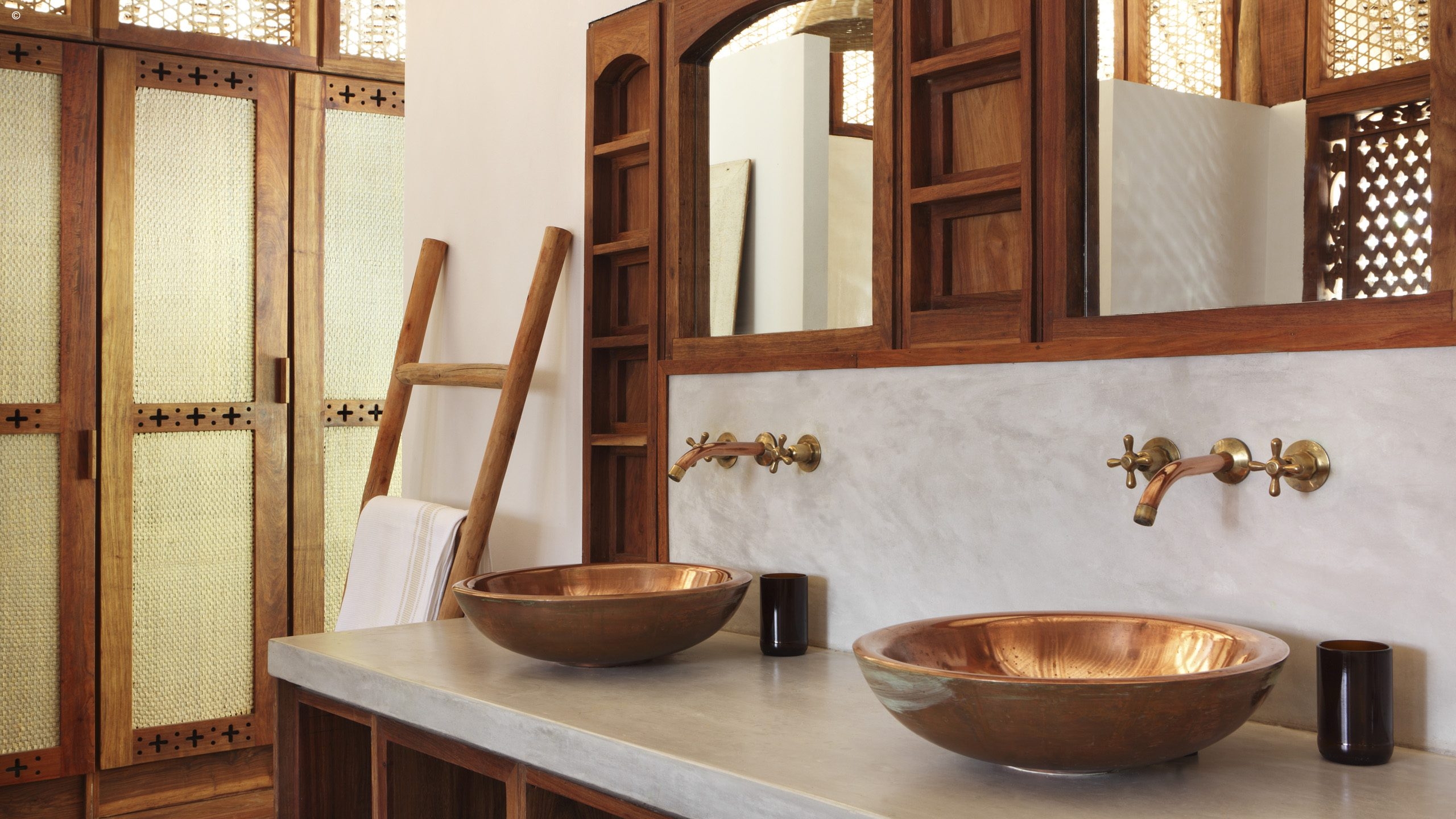 A stylish bathroom featuring two copper sinks, brass faucets, and a wooden ladder, with elegant decorative cabinetry.