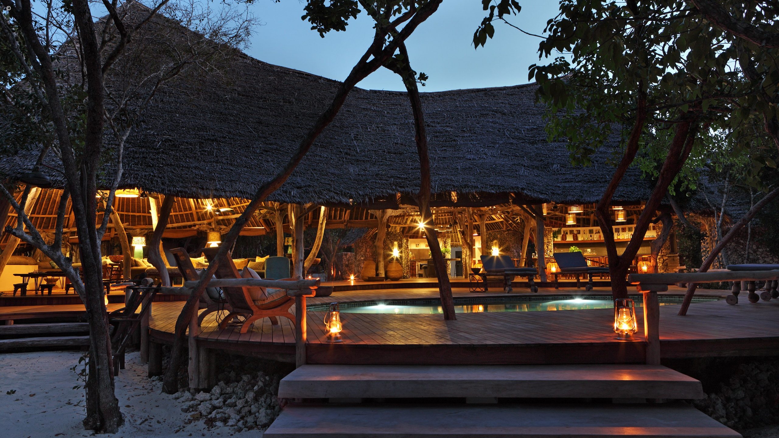 A tropical lodge with a thatched roof, warm lighting, and a wooden deck surrounding a tranquil pool, surrounded by lush trees.