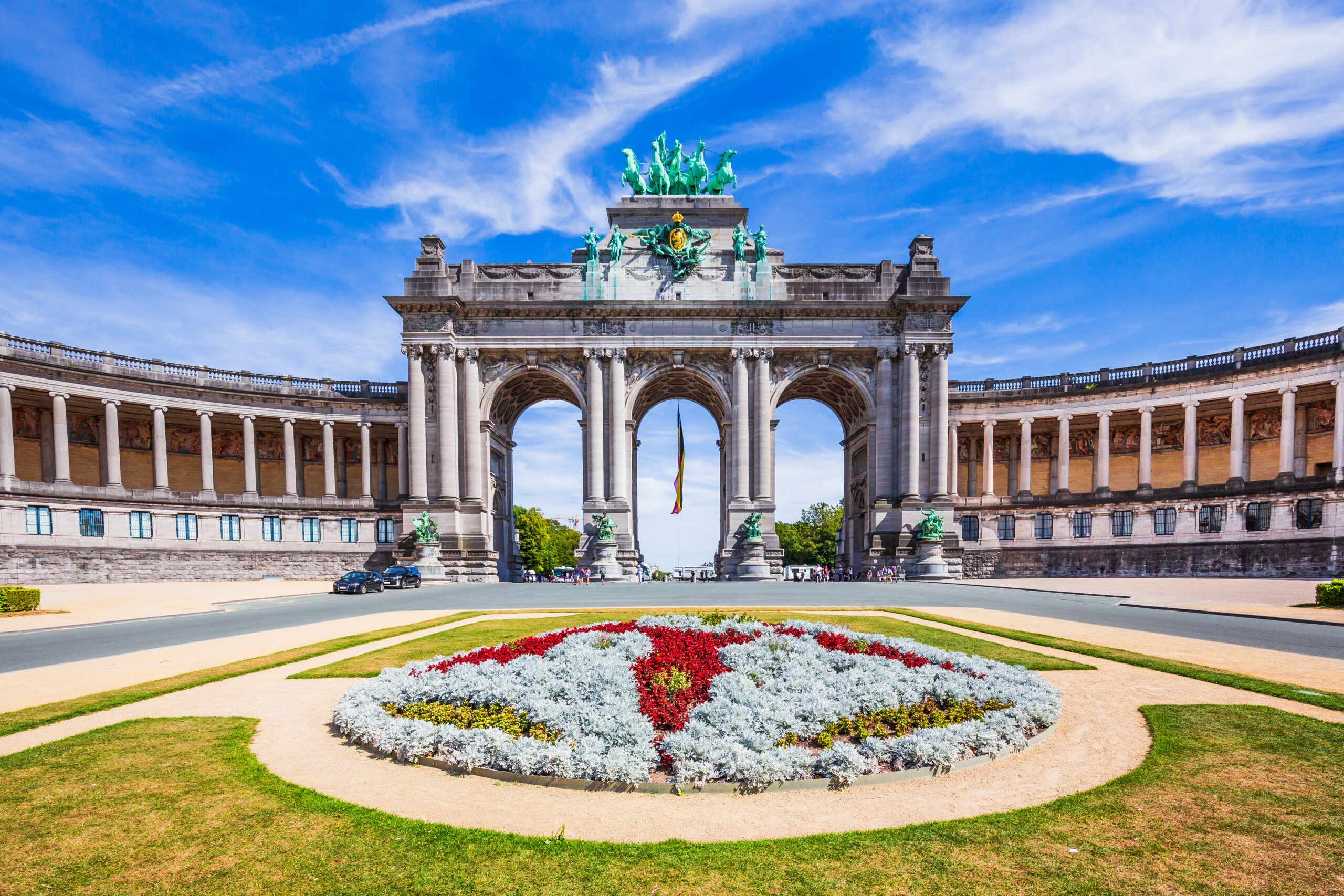 A grand triumphal arch with sculptural details and a colourful floral foreground under a blue sky.