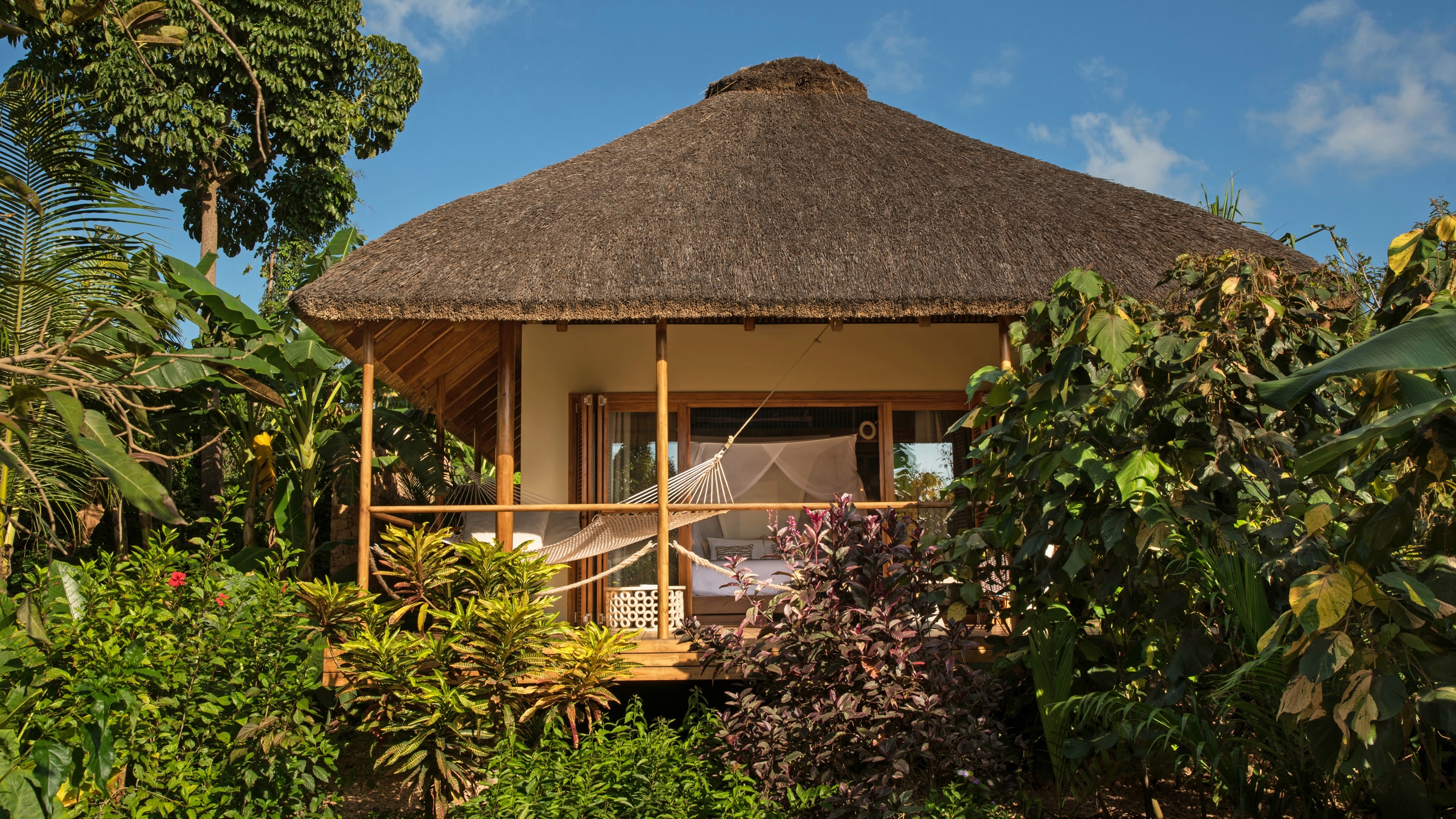 Exterior of a wooden bungalow with a high thatched roof surrounded by lush green and purple foliage.