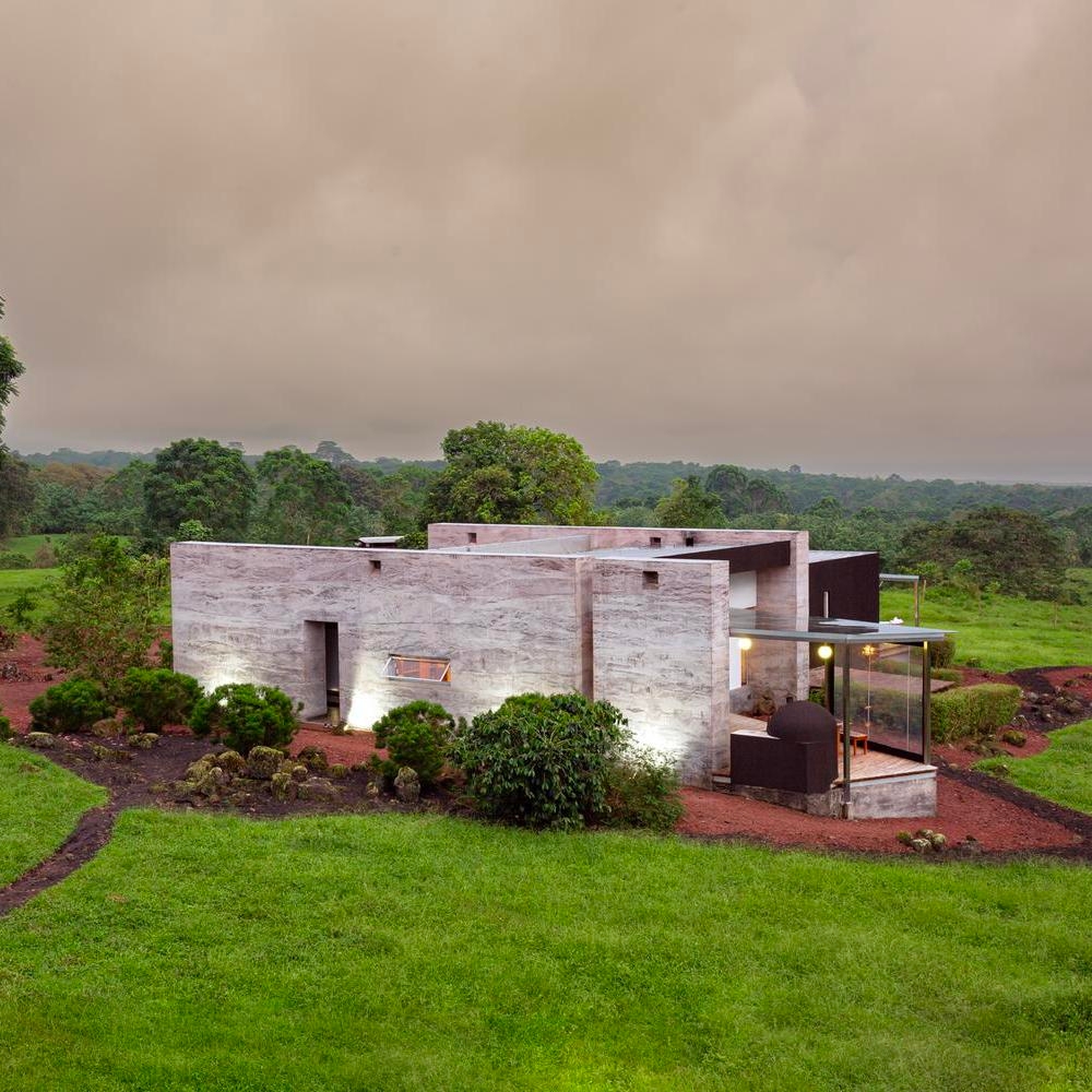 A modern concrete house with a glass room extension, set in a lush green landscape under a cloudy sky.