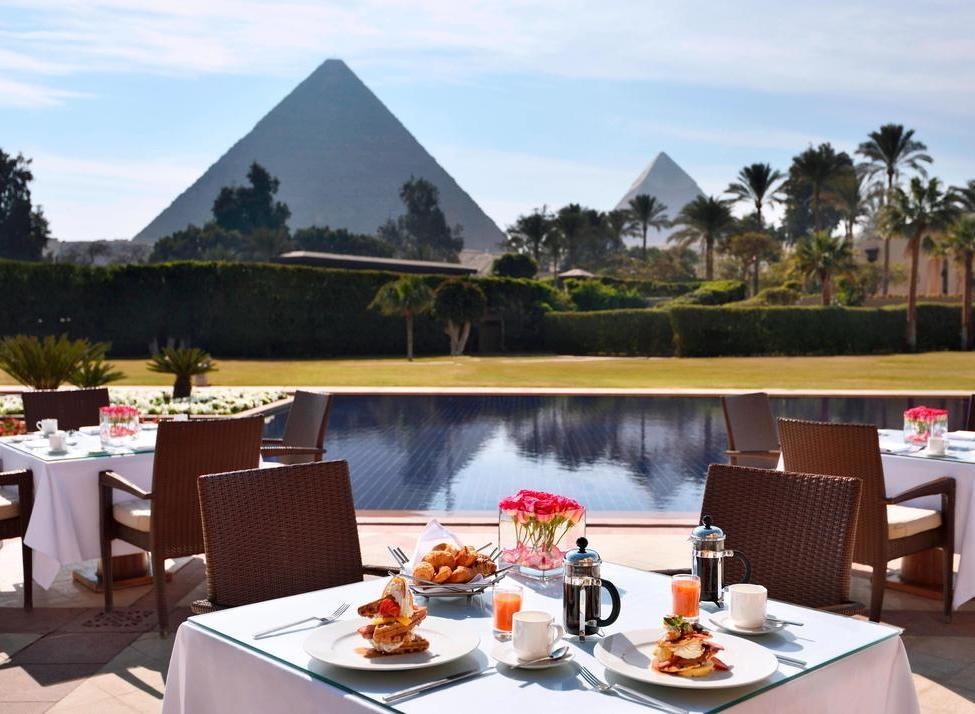 A breakfast table with pastries and coffee overlooks a swimming pool with the Giza Pyramids in the background.