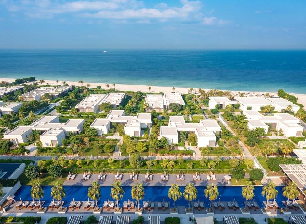 Aerial shot of luxury white villas and a long swimming pool next to a sandy beach and blue ocean.