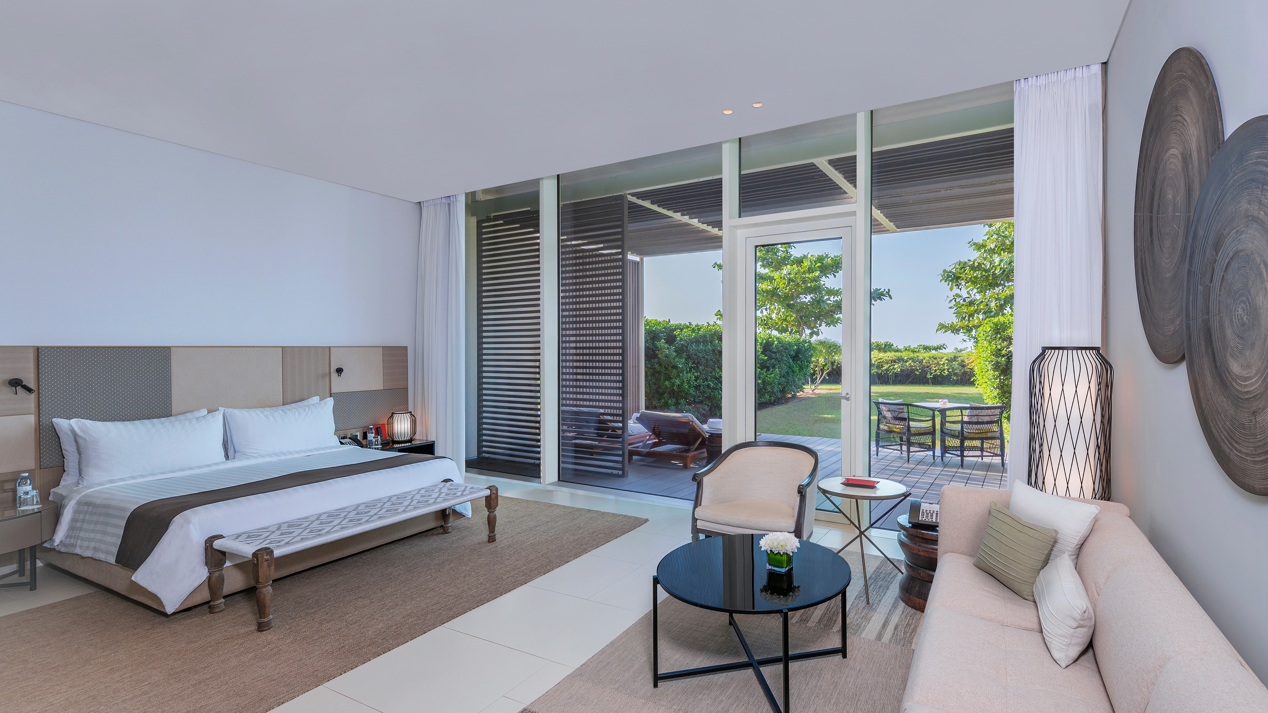 Modern hotel suite with a large bed, tan sofa, and floor-to-ceiling windows leading to a lush private garden.