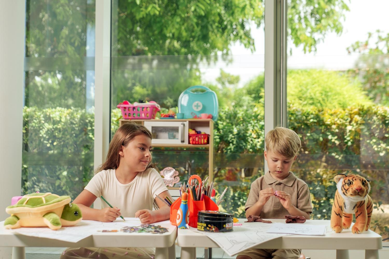 A girl and boy sitting at white tables playing with clay and pencils in a bright room with large windows.