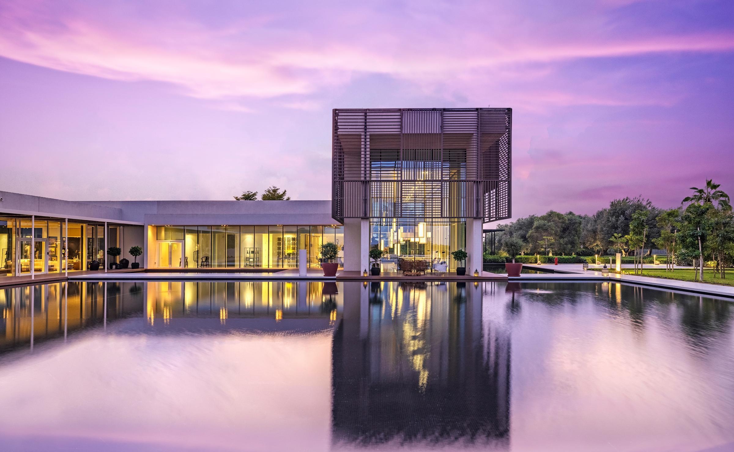 Modern resort architecture with a slatted wood exterior illuminated at dusk above a large, calm reflecting pool.