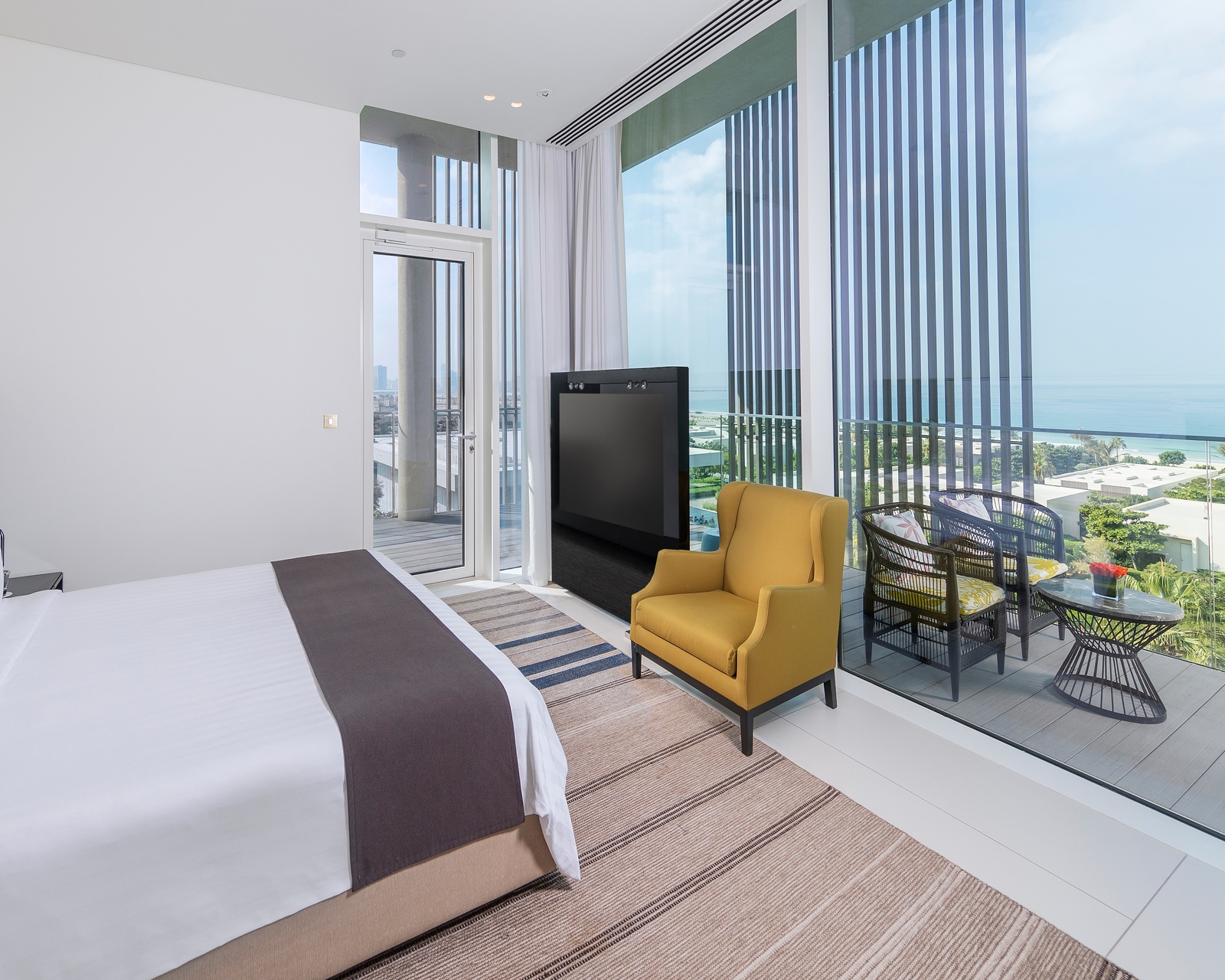 Luxury bedroom with white walls, a yellow chair, and large windows showing a balcony and ocean view.