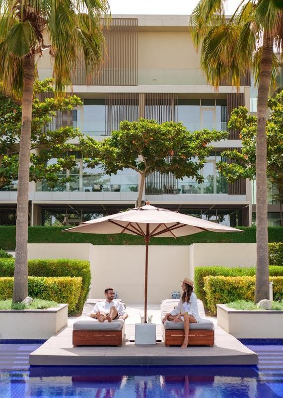 A man and woman sit on lounge chairs under a white umbrella between palm trees by a blue swimming pool.