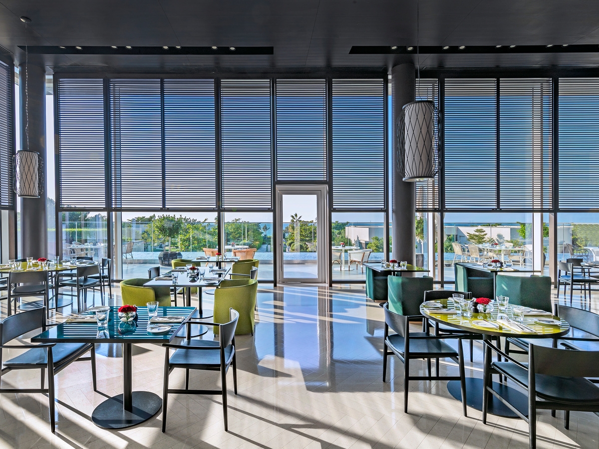 Modern dining room with glass walls, sunlight patterns on the floor, and set tables overlooking the landscape.