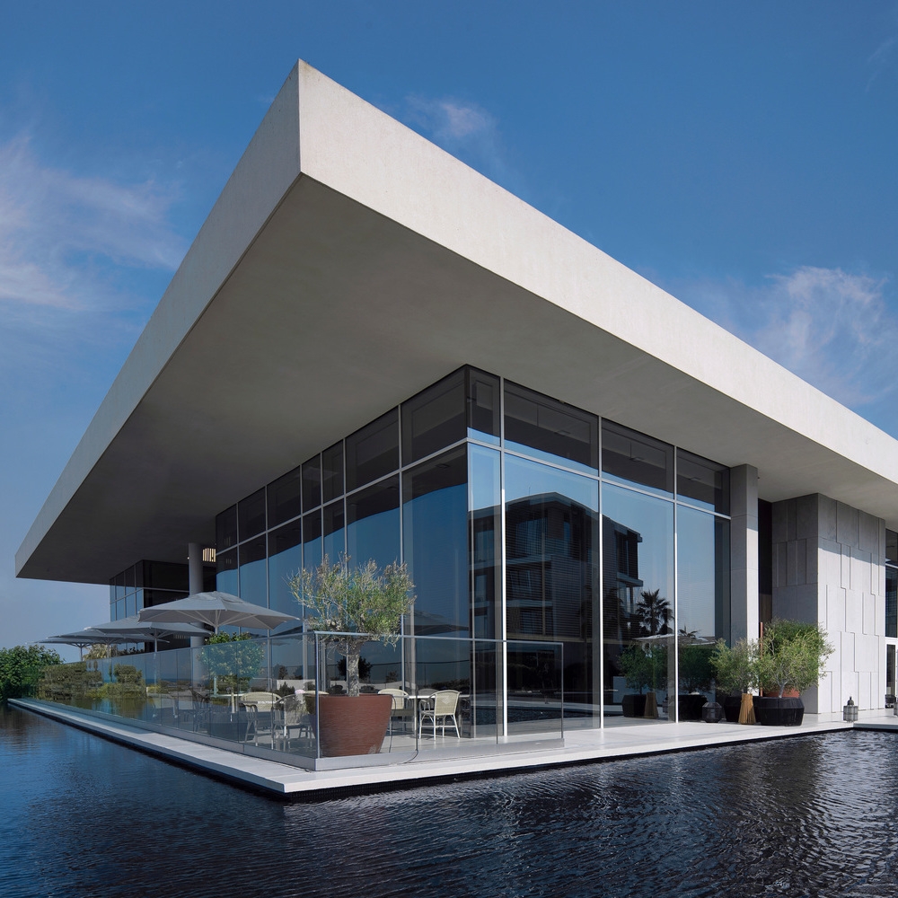 Modern glass building with a large overhanging white roof surrounded by a shallow pool under a blue sky.