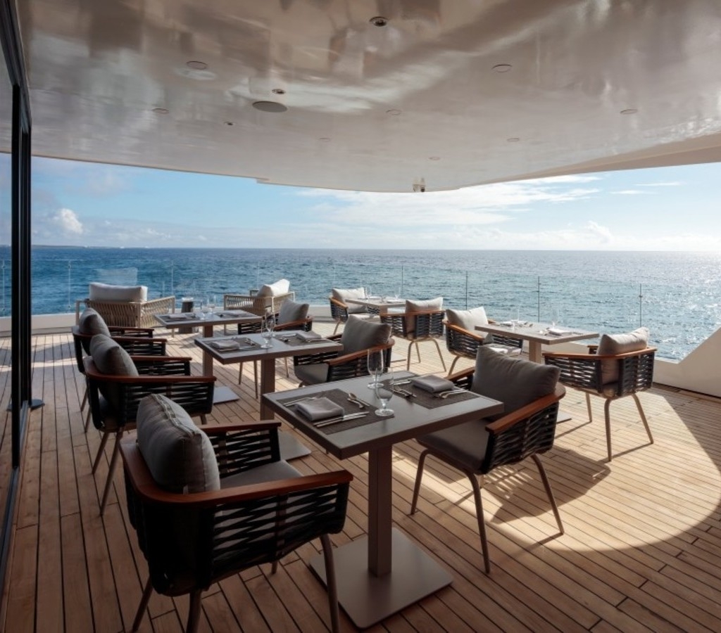 Covered wooden deck of a catamaran set with dining tables and chairs facing the ocean.