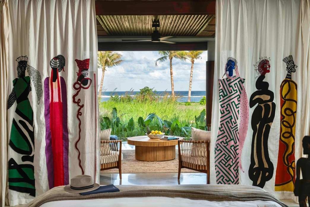 View from a bedroom at Cap Karoso through illustrated curtains showing an outdoor lounge area, palm trees, and the sea.