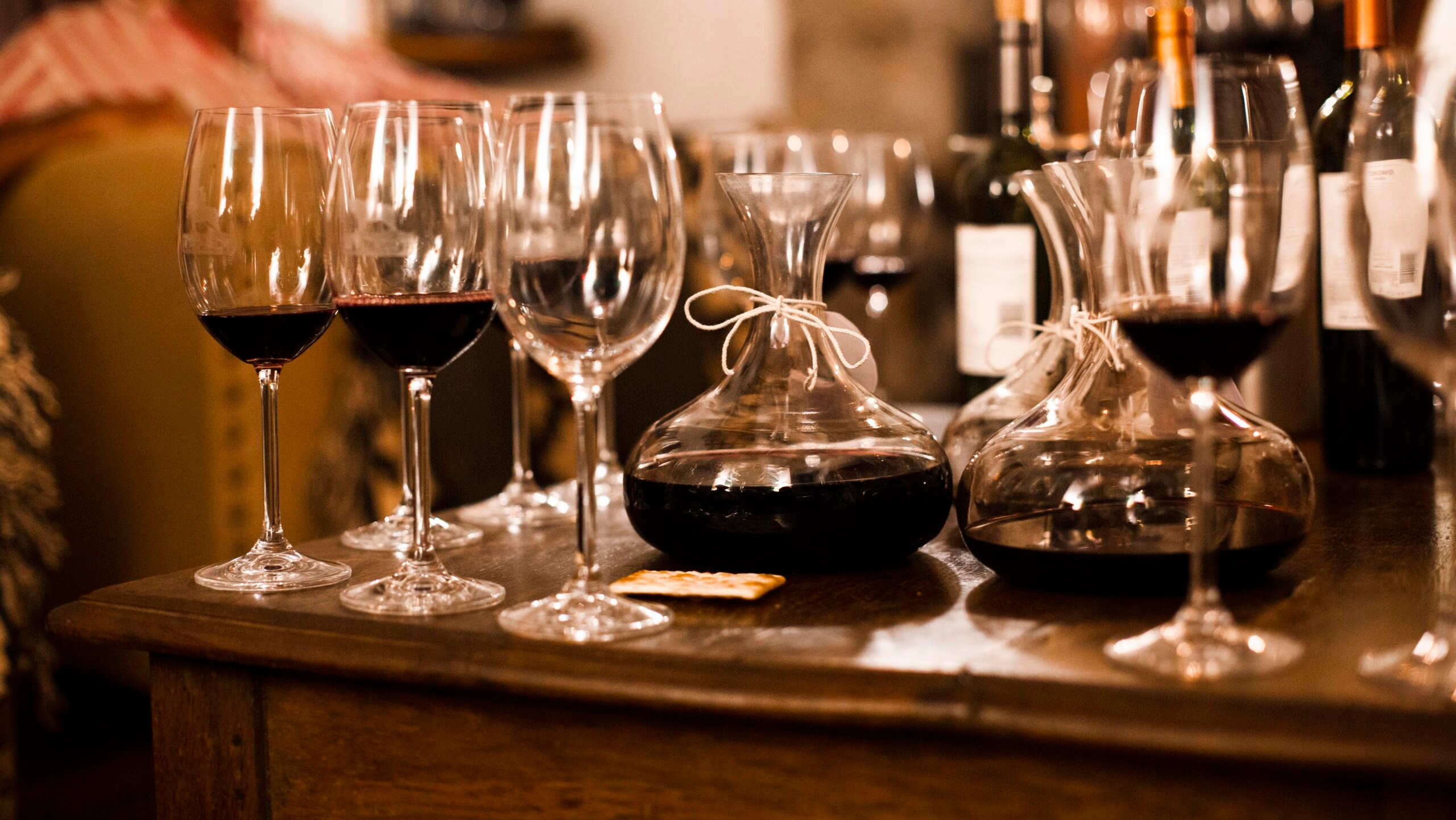 Red wine in glasses and decanters on a wooden table, with bottles in the background.