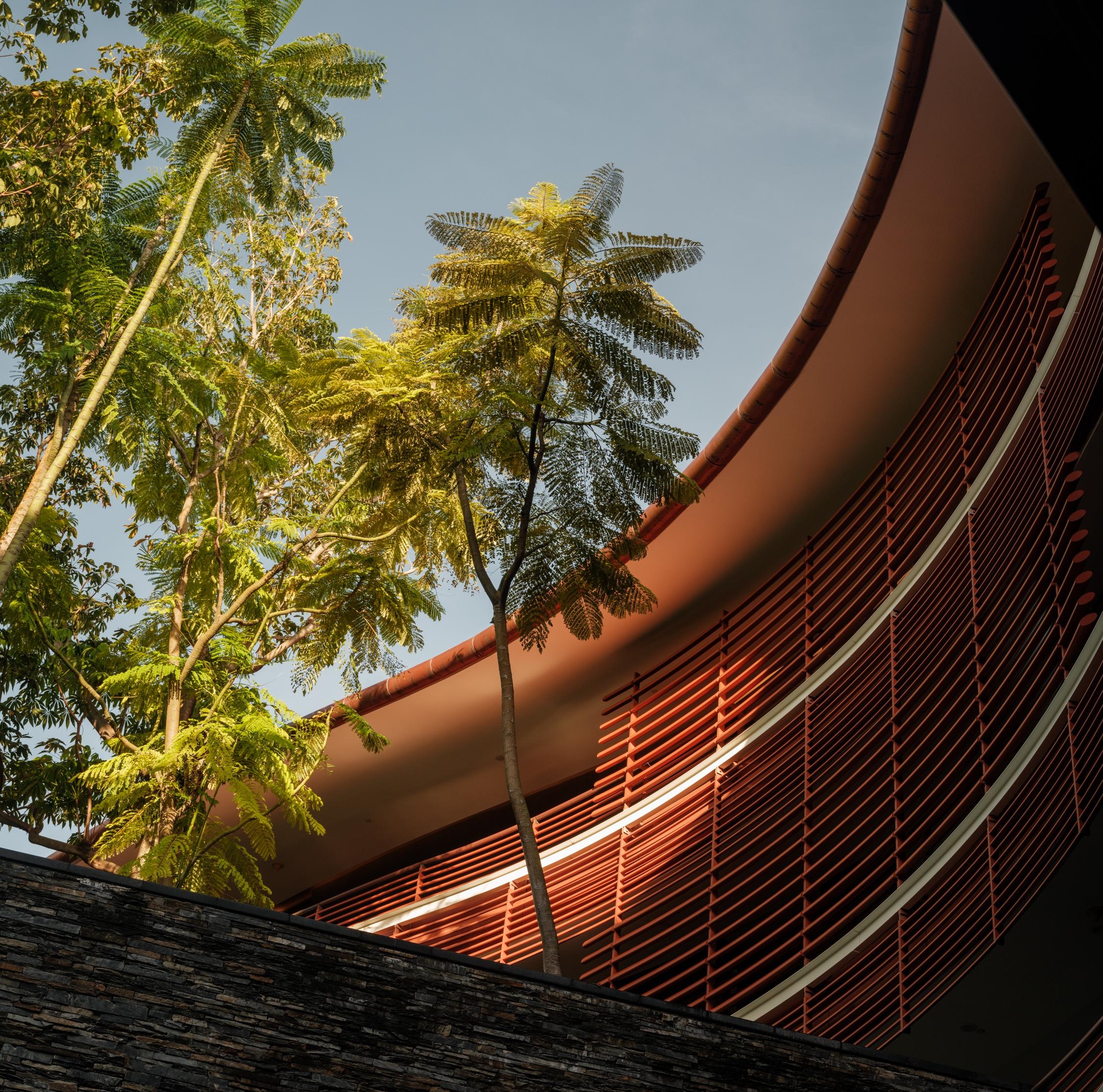 Lush trees in front of a curved building with horizontal red louvers against a clear sky.
