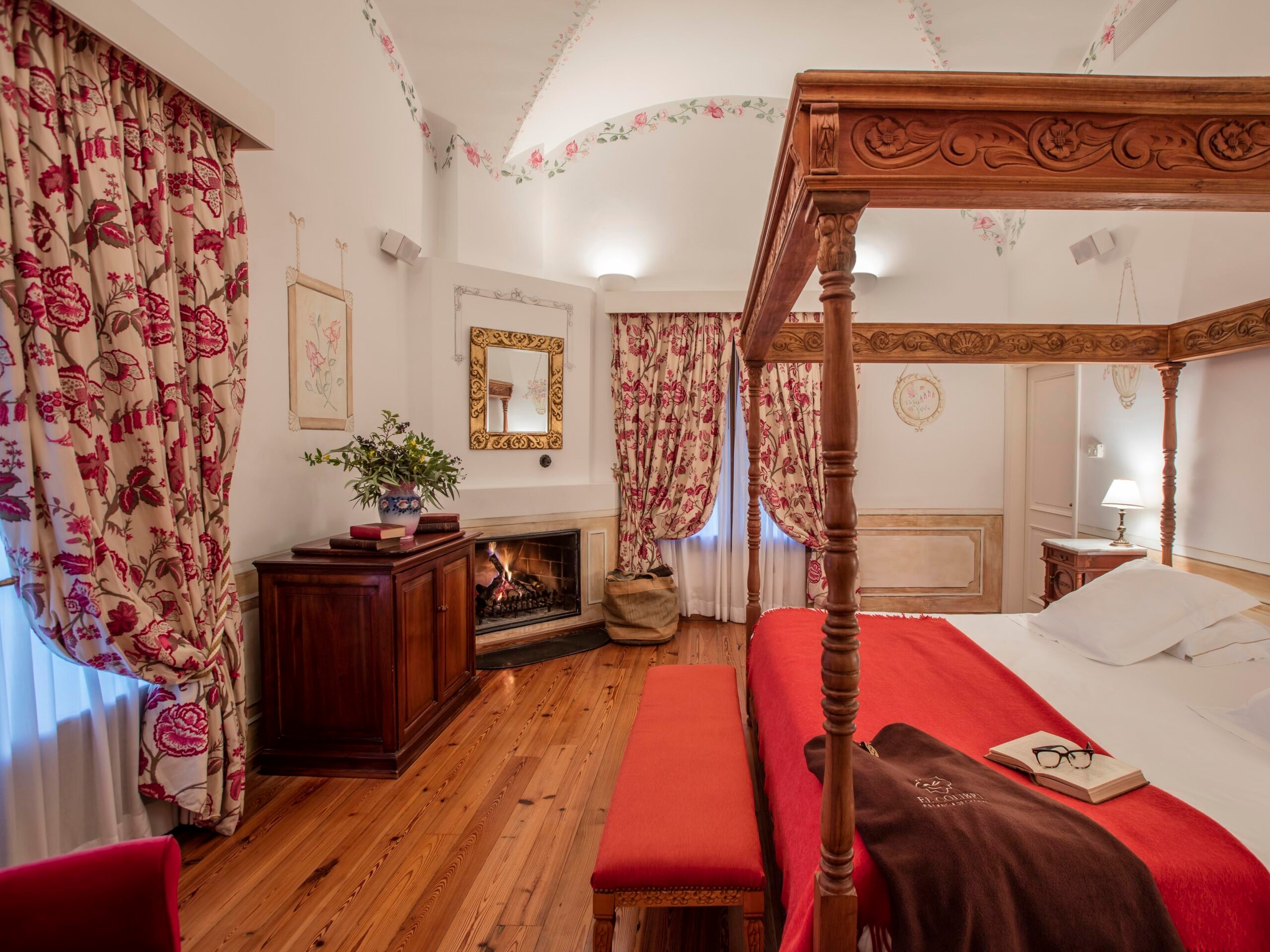 Elegant bedroom with four-poster bed, red accents, fireplace, and floral drapes.