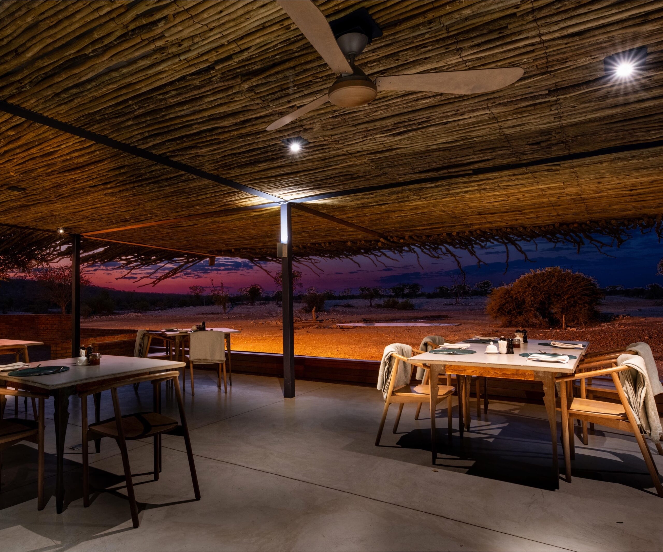 Open-air dining area with tables under a thatched roof at dusk, overlooking a serene, dusky landscape.